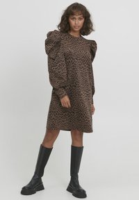 Robe en imprimé léopard dans des tons de marron avec des manches bouffantes, un design mi-long et une texture lisse. Portée avec des bottes noires hautes.