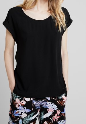 Kobieta w czarnej bluzce z krótkim rękawem, z podwiniętymi rękawami, i spodniach z tropikalnym kwiatowym wzorem, z rękami w kieszeniach, na jednolitym tle.