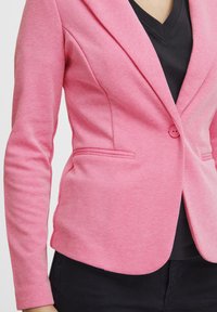 Blazer rosa testurizzato con un design aderente, chiusura a un bottone e due tasche frontali. Indossato sopra un top Nero a V.