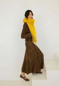 Braune Wildlederjacke, langer brauner Rock mit vertikalen Nähten, leuchtend gelber Schal und Ballerinas im Leopardenmuster. Strukturierte Stoffe und geschichtetes Design.