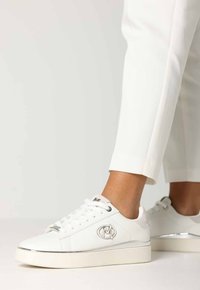 Bílé kožené tenisky s lesklým stříbrným lemem a detailním logem, plochou podrážkou a šněrovacím designem. V kombinaci s bílými zúženými kalhotami.