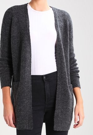 Femme portant un cardigan gris foncé ouvert sur un haut blanc côtelé et un jean noir taille haute sur un fond uni.
