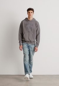 Hanorac gri cu logo "BOSS", buzunar tip cangur, asortat cu blugi desți de nuanță albastră și adidași albi. Design simplu pe un fundal neutru.