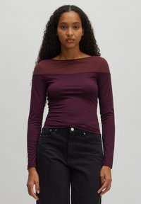 Langärmliges Oberteil aus tiefviolettem Stoff mit durchsichtigem Ausschnitt; taillierte Passform. Kombiniert mit hochgeschnittenen, dunklen Denim-Jeans.