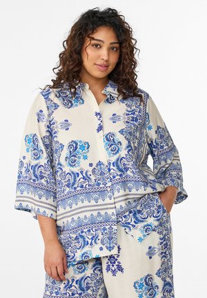 Lokkis juustega naine, kes kannab kreemjat kostüümi siniste keerukate lille- ja paisleimustritega, poseerib ühe käega taskus valge tausta ees.