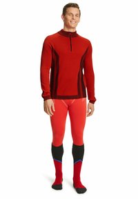 Pull rouge à manches longues avec un zip quart et des accents verticaux noirs, associé à un legging rouge moulant et des chaussettes noires hautes avec des détails rouges et bleus.