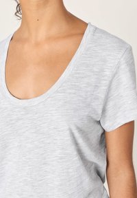 Artimas kadro vaizdas, kuriame matyti asmuo dėvintis šviesiai pilką trumpomis rankovėmis marškinėlį su apvalia iškirpte, matomi kaklas, raktikaulis ir viršutinė krūtinės dalis.