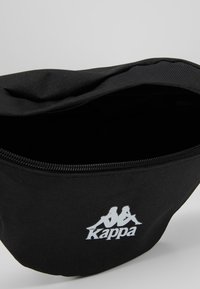 Czarna nerka Kappa z teksturowaną powierzchnią, zamknięciem na zamek błyskawiczny u góry oraz białym logo z przodu. Wnętrze jest pojemne i niepodszewkowane.