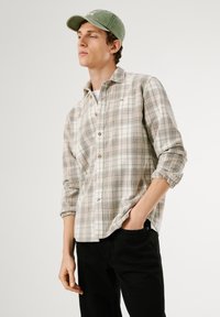 Camicia a quadri chiara nei toni del beige e dell'olive, con chiusura a bottoni, maniche arrotolabili e taschino sul petto. Abbinata a pantaloni neri.