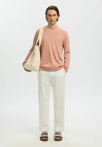 Jeune homme portant un pull rose clair, un pantalon crème, des sandales beige, tenant un grand sac fourre-tout beige sur une épaule, debout devant un fond uni.