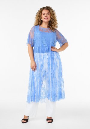 Vrouw met krullend haar staat rechtop in een doorschijnende blauwe kanten jurk over een blauwe top en witte wijde broek, met zwarte sandalen met hak.
