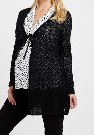 Femme enceinte portant un chemisier blanc à taches noires sous un cardigan en dentelle noire à motif, montrée des épaules jusqu'à mi-cuisse.
