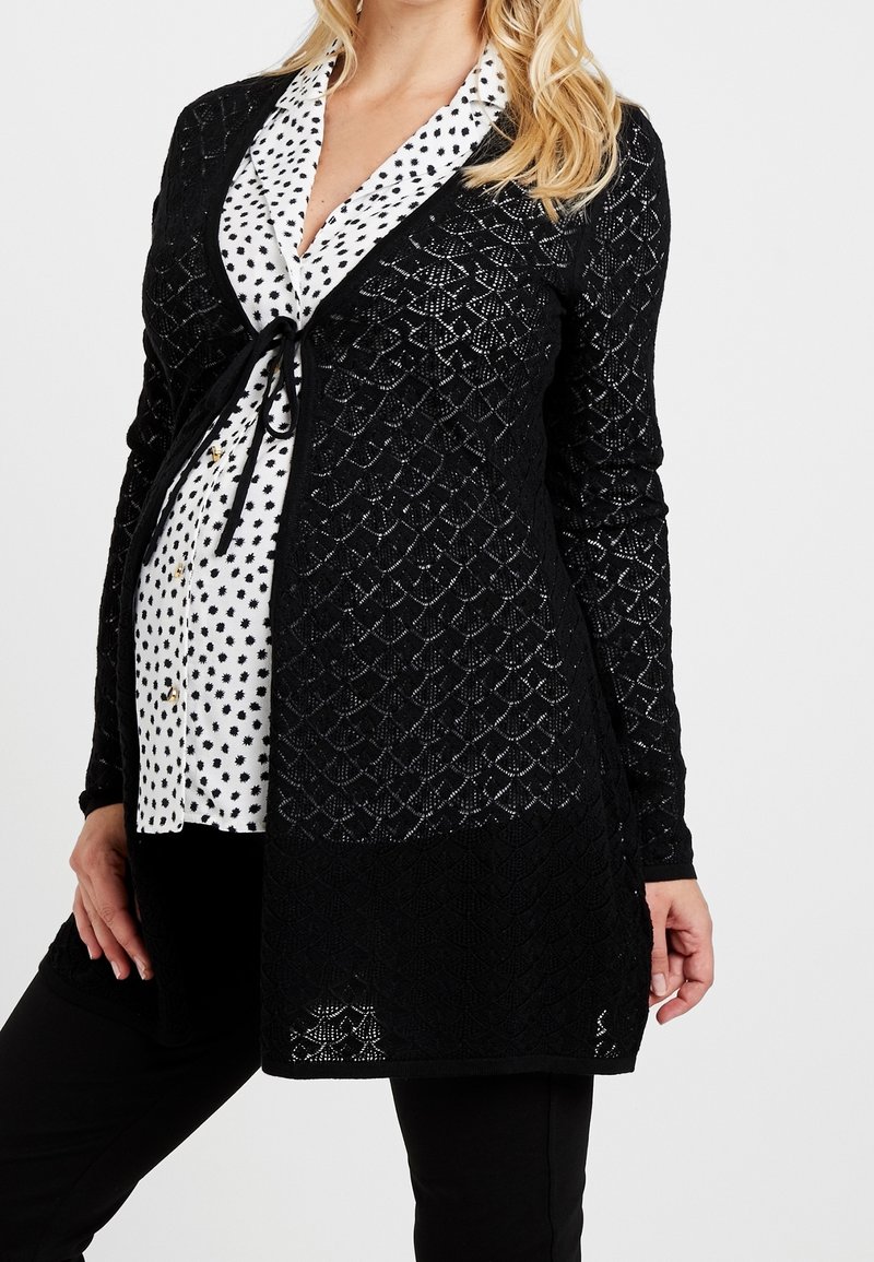 Femme enceinte portant un chemisier blanc à taches noires sous un cardigan en dentelle noire à motif, montrée des épaules jusqu'à mi-cuisse.