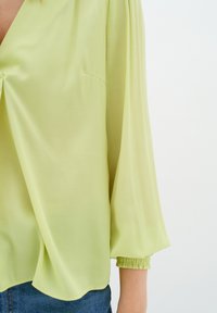 Blusa verde lime con scollatura a V profonda, maniche a sbuffo a tre quarti e polsini arricciati, realizzata in tessuto leggero e liscio.