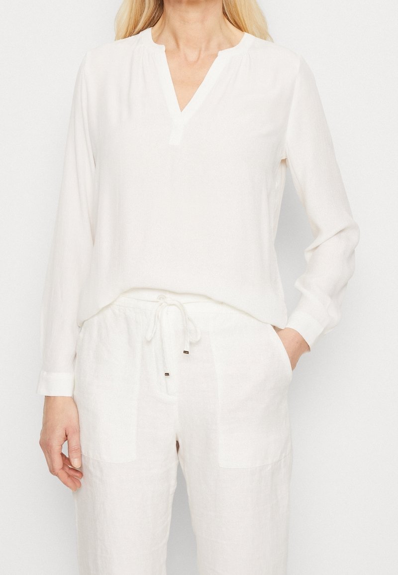 Haut blanc à manches longues avec un col en V et une coupe décontractée. Fabriqué dans un tissu léger. Associé à un pantalon à cordon assorti.