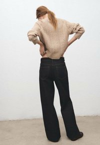 Suéter de punto beige con patrón texturizado, combinado con pantalones vaqueros de pierna ancha negros de tiro alto, que muestran un dobladillo deshilachado y detalles de costura oscura.