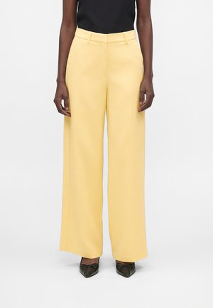 Person iført blegegule bukser med høj talje og vide ben med bæltestropper, kombineret med sorte spidse sko og en sort ærmeløs top.