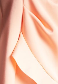 Světlý broskvový materiál s jemnou texturou, který obsahuje drapované části a mírný lesk. Okraje jsou jemně zakřivené s čistým prošitím.