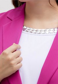 Fuchsia blazer med strukturerade slag, i kombination med en vit topp med en rad av reflekterande stenar längs kragen. En hand rör vid blazern.