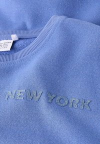 Felpa azzurro chiaro con tessuto strutturato. "NEW YORK" ricamato con lettere luccicanti sul petto. Design con scollo rotondo, tessuto morbido.