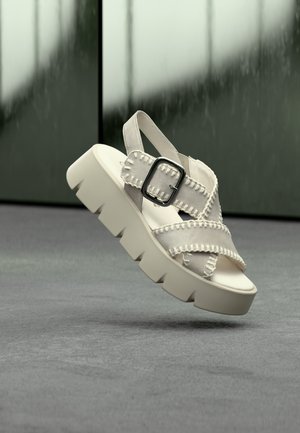 Witte platformsandaal met een breed bandontwerp, voorzien van een gesp en decoratieve stiksels op suède materiaal. Dikke, gestructureerde zool.