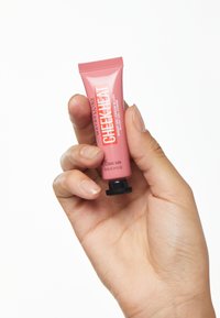 Maybelline Cheek Heat, un piccolo tubetto rosa con un tappo a vite nero, etichettato "15 Nude Burn", tenuto in mano su uno sfondo bianco.