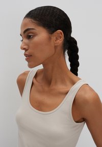 Hellbeiges Tanktop mit rundem Ausschnitt und dünnen Trägern aus glattem Stoff, seitlich an einer Frau gezeigt.