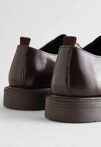 Zapatos de cuero marrón con un acabado brillante, que presentan un tacón bajo y un tirador texturizado, con costuras detalladas a lo largo de las juntas.