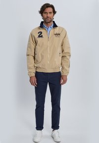 Chaqueta cremosa con cremallera y cuello marino, con un logo, el número "2" y bolsillos laterales. Combinada con pantalones marinos y zapatillas blancas.