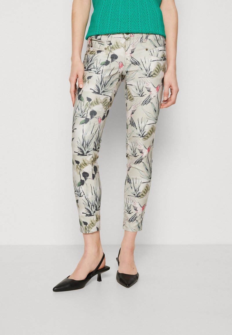 Pantalon slim avec un fond beige et un motif coloré d'oiseaux et de feuillage, porté avec des talons slingback noirs et un haut vert.