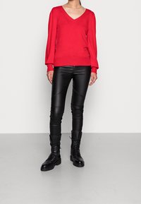 Pull rouge à col en V et manches longues, associé à un pantalon noir en similicuir avec des coutures diagonales et des bottes de combat noires à lacets.