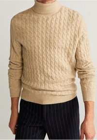 Pull à col roulé beige en maille torsadée porté avec un pantalon rayé bleu marine foncé sur une personne debout devant un fond uni.