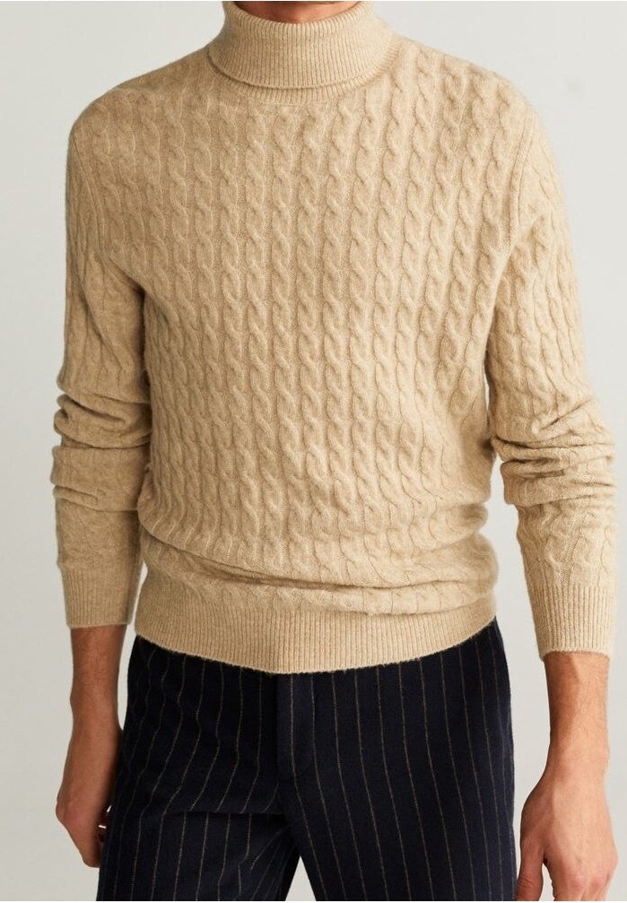 Pull à col roulé beige en maille torsadée porté avec un pantalon rayé bleu marine foncé sur une personne debout devant un fond uni.