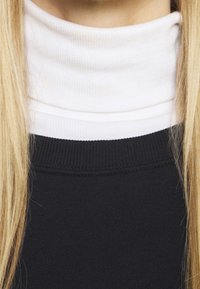 Suéter negro con un dobladillo texturizado, superpuesto a un cuello alto blanco con canalé. La tela parece suave, con un ajuste ceñido alrededor del cuello.