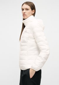 Γυναίκα που φοράει λευκό μπουφάν με επένδυση, ψηλό γιακά και κουκούλα, στέκεται με το ένα χέρι στην τσέπη των μαύρων παντελονιών, μπροστά σε απλό φόντο.