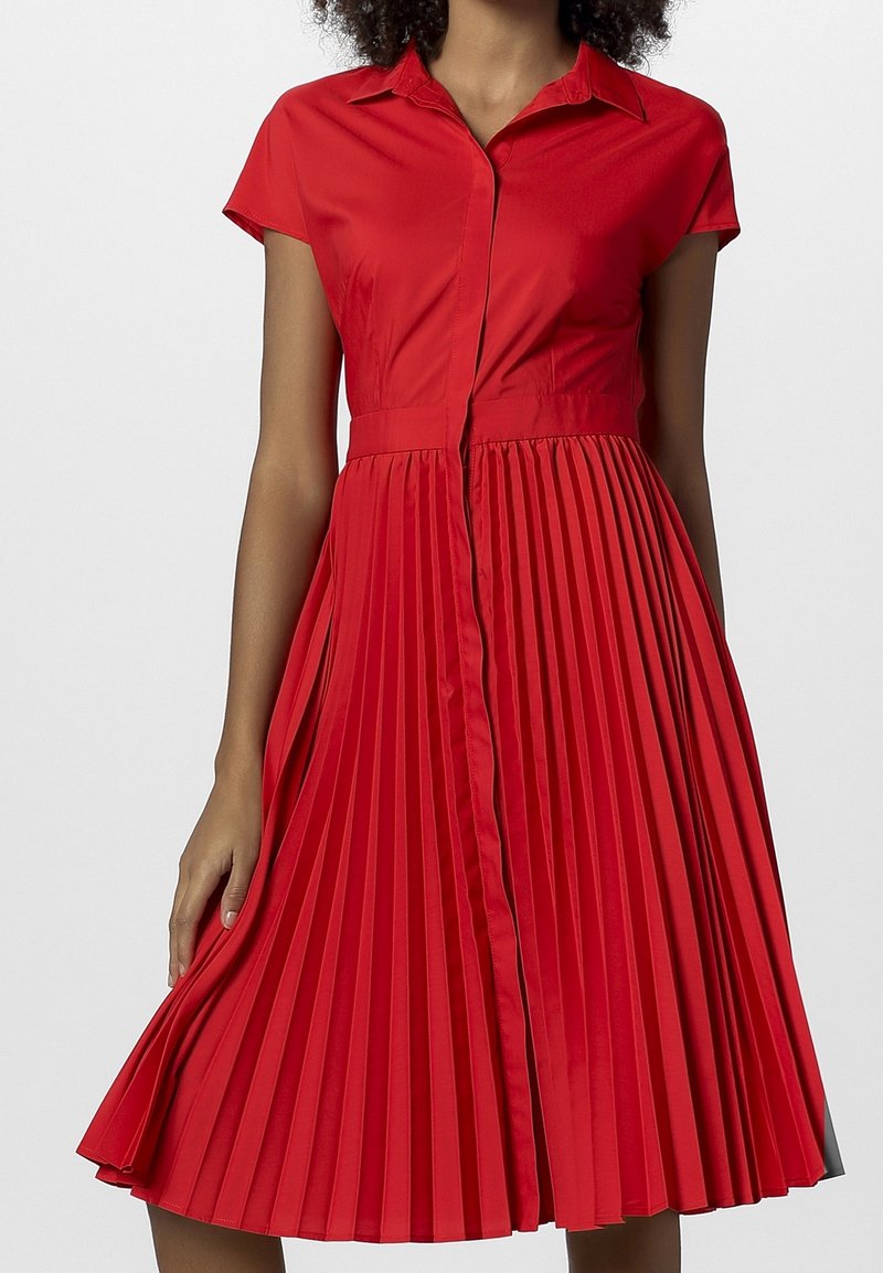 Femme portant une robe rouge à manches courtes avec col, taille cintrée et jupe plissée, debout devant un fond uni.