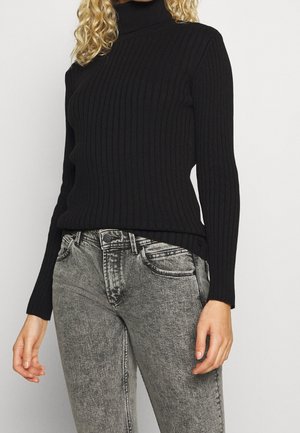 Nainen, jolla on musta ribbineuleinen pooloneule ja korkeavyötäröiset haalistuneet harmaat farkut, seisoo yksinkertaisen taustan edessä.