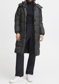 Abrigo acolchado largo negro con capucha, que cuenta con un patrón acolchado, cierre frontal con cremallera, bolsillos laterales y puños elásticos. Combinado con pantalones negros y zapatillas blancas.