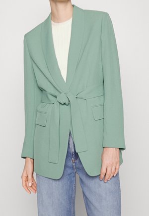 Model iført en lysegrøn blazer med bælte over en hvid ribbet top og lyseblå jeans, hænderne afslappede langs siderne.