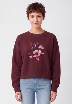 Jeune femme aux cheveux courts et bruns portant un sweat-shirt bordeaux avec un motif floral rose et bleu et un jean bleu clair, debout sur fond blanc.