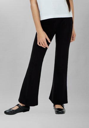 Femme portant un pantalon noir à jambes larges, des chaussures Mary Jane noires et un haut blanc, debout devant un fond gris uni.