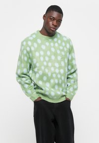 Groen gebreid sweater met een patroon van lichtblauwe stippen. Gemaakt van pluizig breimateriaal met een geribbelde kraag en manchetten. Draagt in combinatie met zwarte broek.