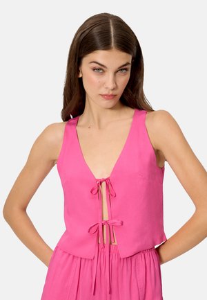 Mujer vestida con una blusa sin mangas de color rosa con dos lazos frontales y una falda rosa a juego de cintura alta, de pie con las manos en las caderas contra un fondo liso.