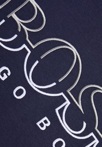 Marinblått tyg med stor, intrikat broderad text i vitt och ljusgrått, som visar en slät textur och distinkta bokstavsformer.