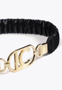 Schwarzes elastisches Haargummi mit einem goldenen Metallelement, das sich überlappenden Buchstaben zeigt. Glatte Textur mit gerafften Details im elastischen Band.