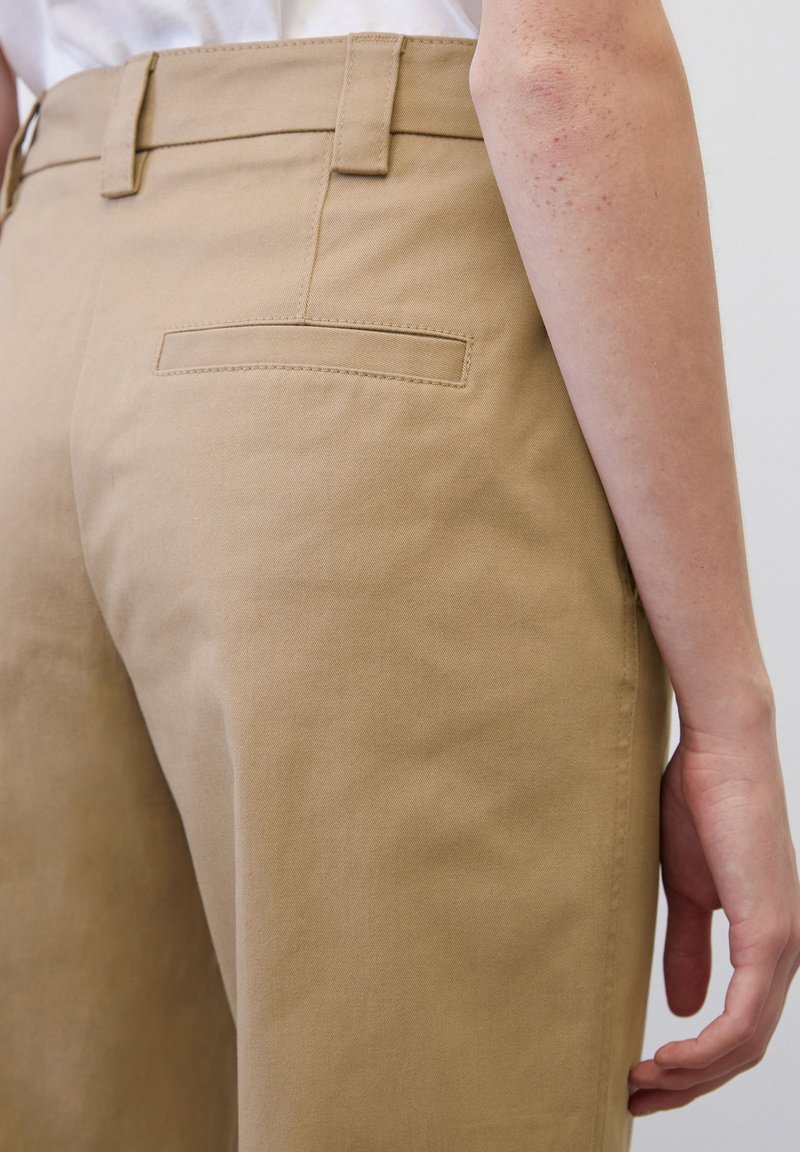 Béžové bavlněné kalhoty s hladkou texturou, s jednou zadní kapsou a poutky na opasek, zobrazené z bočního úhlu.