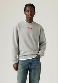 Sweat-shirt gris en tissu mélangé coton avec un col rond, arborant un logo rouge de Levi's sur la poitrine, associé à un jean en denim bleu.