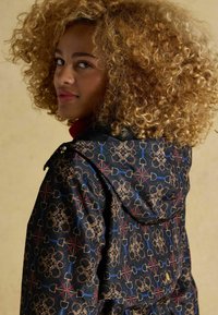 Melnā rakstainā jaka ar ziedu un ģeometriskajiem dizainiem zelta, sarkanā un zilā akcentos. Ar kapuci un teksturētu apkakli.