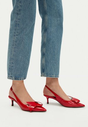 Persona con jeans azules recortados y tacones slingback rojos puntiagudos con tacones bajos tipo gatito y detalle de lazo.