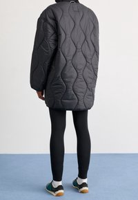 Chaqueta acolchada negra de plumas con un patrón ondulado, de corte holgado, cuello alto y hombros caídos, llevada sobre leggings negros y zapatillas.